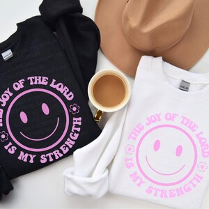 May include: Black and white sweatshirts with the text "THE JOY OF THE LORD IS MY STRENGTH" in pink, featuring a smiley face design. A tan hat and a cup of coffee are also in the image.