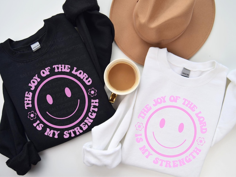 May include: Black and white sweatshirts with the text "THE JOY OF THE LORD IS MY STRENGTH" in pink, featuring a smiley face design. A tan hat and a cup of coffee are also in the image.