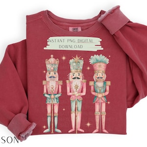 May include: A red sweatshirt with a graphic of three nutcracker soldiers. The nutcrackers are wearing pink and blue uniforms and holding swords. The text "INSTANT PNG DIGITAL DOWNLOAD" is printed on the sweatshirt.