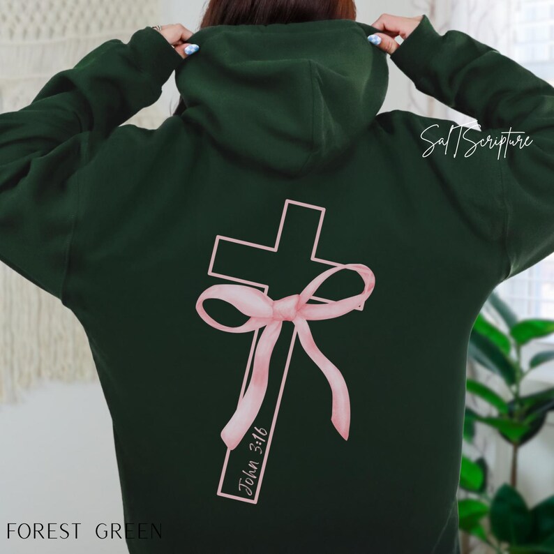 May include: Forest green hoodie with a pink cross and bow design on the back. The text "John 3:16" is printed on the cross. The hoodie has a hood and long sleeves.