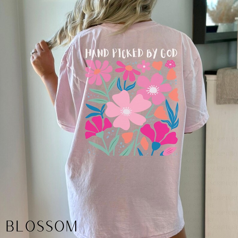 May include: A light pink t-shirt with a floral design on the back. The design features pink, fuchsia, and orange flowers with green and blue leaves. The text "HAND PICKED BY GOD" is above the floral design. The word "BLOSSOM" is at the bottom.