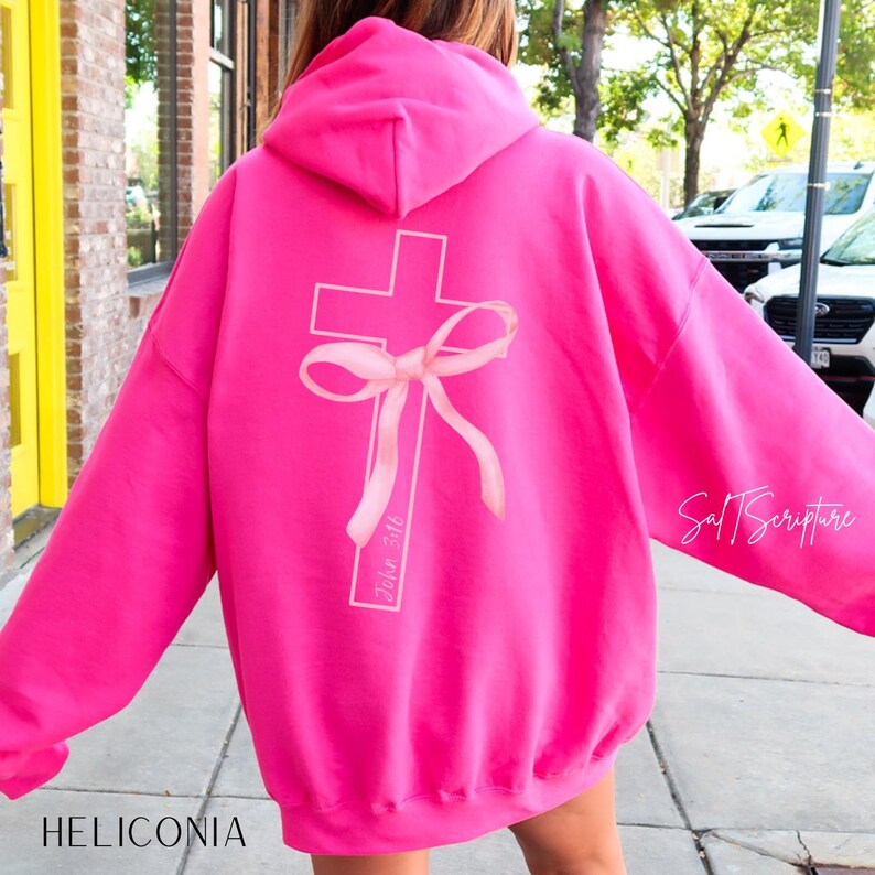 May include: A bright pink hoodie with a white cross design and a pink bow on the back. The text "John 3:16" is printed vertically on the cross. The brand name "Salt Scripture" is printed on the right sleeve.