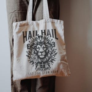 May include: A beige tote bag with a black and white graphic design featuring a lion's head with a crown and the text "Hail Hail Lion of Judah Let the Lion Roar".