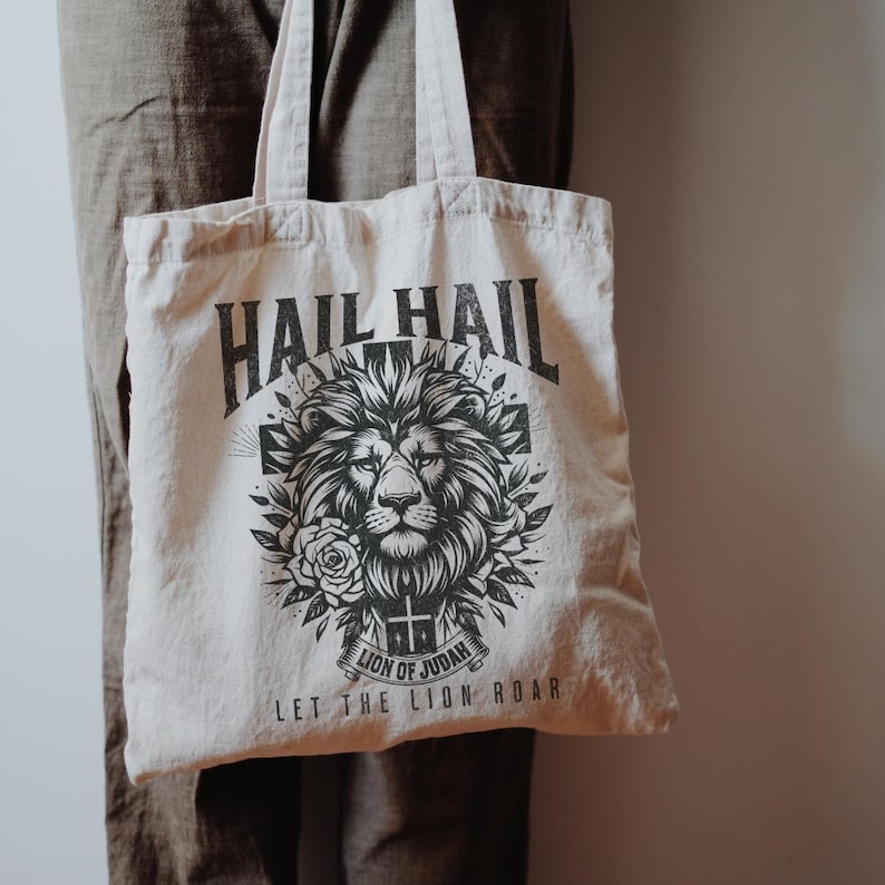 May include: A beige tote bag with a black and white graphic design featuring a lion's head with a crown and the text "Hail Hail Lion of Judah Let the Lion Roar".