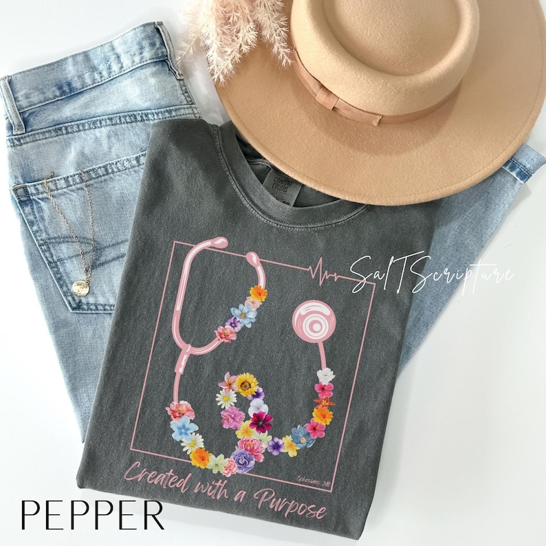 May include: A dark gray t-shirt featuring a pink stethoscope design adorned with colorful flowers. The shirt includes the text "Created with a Purpose" and is paired with light blue jeans and a tan hat. The word "PEPPER" is at the bottom.