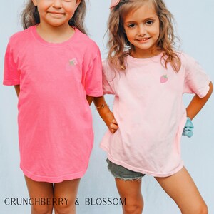May include: Two children wearing pink and light pink t-shirts with embroidered strawberry designs. The girl on the left is wearing a bright pink t-shirt with a strawberry and leaves design. The girl on the right is wearing a light pink t-shirt with a single strawberry design.  "Crunchberry & Blossom" is written at the bottom of the image.