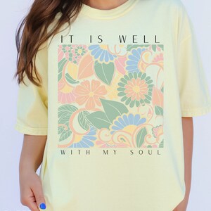May include: A yellow t-shirt with a floral design and the text "It is well with my soul".