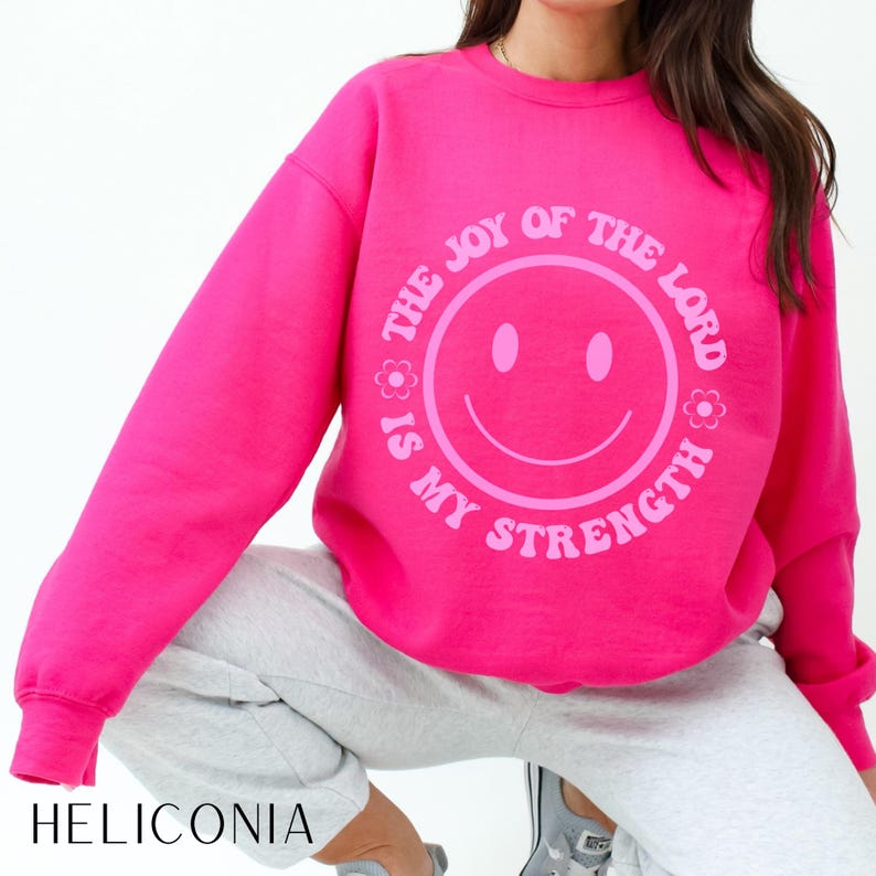 May include: A bright pink sweatshirt with the text "THE JOY OF THE LORD IS MY STRENGTH" encircling a smiley face. The sweatshirt is a solid color and has long sleeves. The word "HELICONIA" is printed at the bottom.