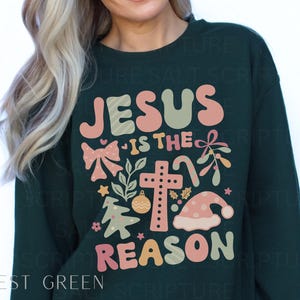 May include: Forest green sweatshirt with a Christmas-themed graphic that says "Jesus is the reason" with a cross, a bow, a candy cane, and a Christmas tree.