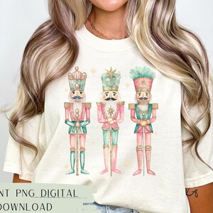 May include: A white t-shirt with a design of three watercolor nutcracker soldiers. The nutcrackers are wearing pink, green, and blue uniforms with gold accents. The design is centered on the shirt.