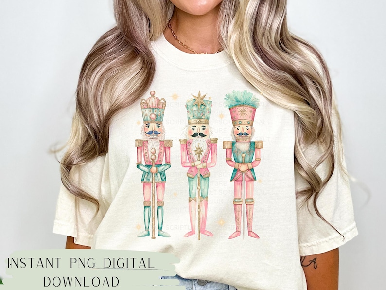 May include: A white t-shirt with a design of three watercolor nutcracker soldiers. The nutcrackers are wearing pink, green, and blue uniforms with gold accents. The design is centered on the shirt.