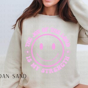 May include: A Gildan Sand sweatshirt with a pink smiley face design. The text around the smiley face reads "The Joy of the Lord is My Strength". The sweatshirt is a neutral color and has long sleeves.