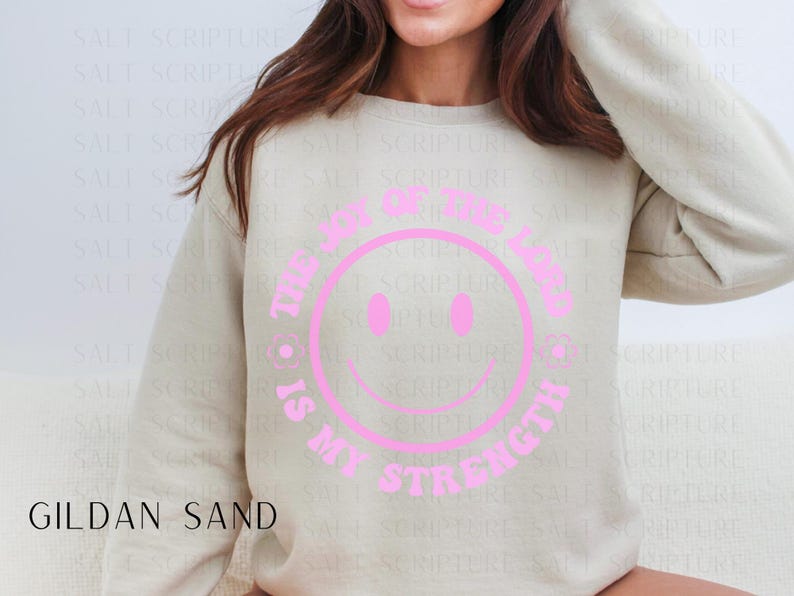 May include: A Gildan Sand sweatshirt with a pink smiley face design. The text around the smiley face reads "The Joy of the Lord is My Strength". The sweatshirt is a neutral color and has long sleeves.
