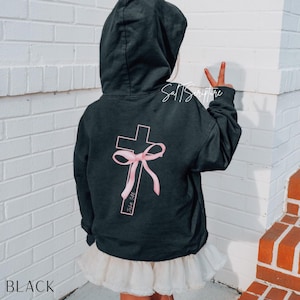 May include: Black hooded sweatshirt with a pink cross design on the back, featuring a bow and the text "John 3:16". The sweatshirt is paired with a white tutu skirt. The word "BLACK" is at the bottom.