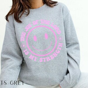 May include: A sports grey sweatshirt with a pink smiley face graphic. The text "THE JOY OF THE LORD IS MY STRENGTH" is printed around the smiley face. The sweatshirt is a casual, comfortable style.