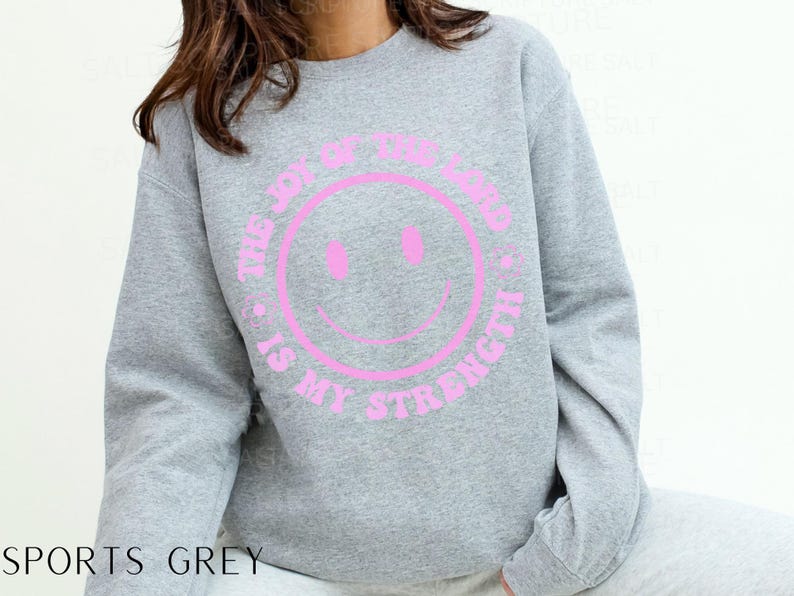 May include: A sports grey sweatshirt with a pink smiley face graphic. The text "THE JOY OF THE LORD IS MY STRENGTH" is printed around the smiley face. The sweatshirt is a casual, comfortable style.