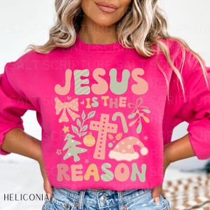 May include: A pink sweatshirt with the text "Jesus is the reason" in a festive font with Christmas-themed illustrations.