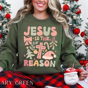 May include: A green Christmas sweatshirt with the text "Jesus is the reason" in pink and white lettering. The design includes a cross, a bow, and a Christmas tree.
