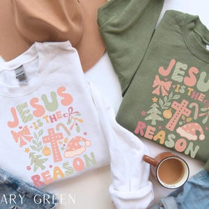 May include: Two Christmas sweatshirts with the text "Jesus is the reason" in a colorful, festive font. The sweatshirts are white and military green.