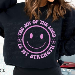 May include: Black sweatshirt with a pink smiley face design. The text around the smiley face reads "The Joy of the Lord is My Strength". The word "BLACK" is printed at the bottom of the sweatshirt.