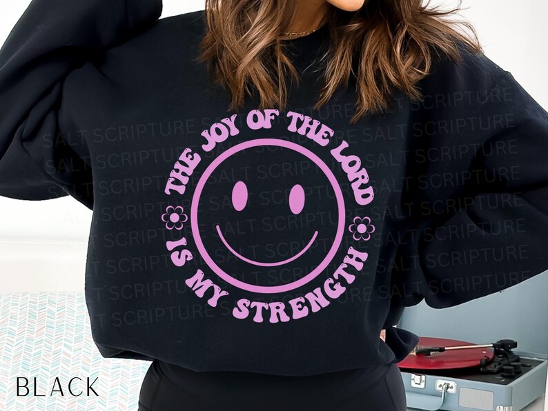 May include: Black sweatshirt with a pink smiley face design. The text around the smiley face reads "The Joy of the Lord is My Strength". The word "BLACK" is printed at the bottom of the sweatshirt.