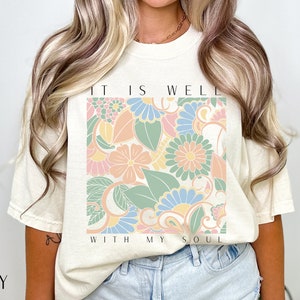 May include: A cream coloured t-shirt with a floral design and the text "It is well with my soul".