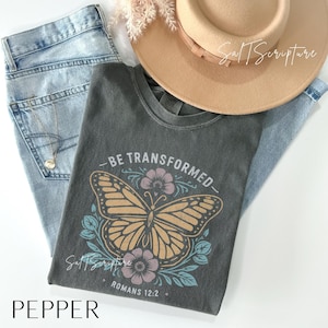 Può includere: Una maglietta grigio scuro con la scritta "BE TRANSFORMED" sopra una grafica a farfalla. La farfalla è circondata da fiori e foglie. Nell'immagine ci sono anche jeans azzurri e un cappello color cachi. La parola "PEPPER" è in basso.