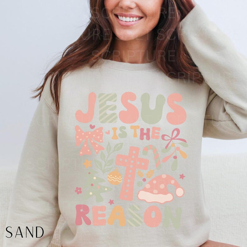 May include: A beige sweatshirt with a Christmas-themed graphic that says "Jesus is the reason" in colorful, stylized letters. The graphic also includes a cross, a bow, candy canes, and a Christmas tree.