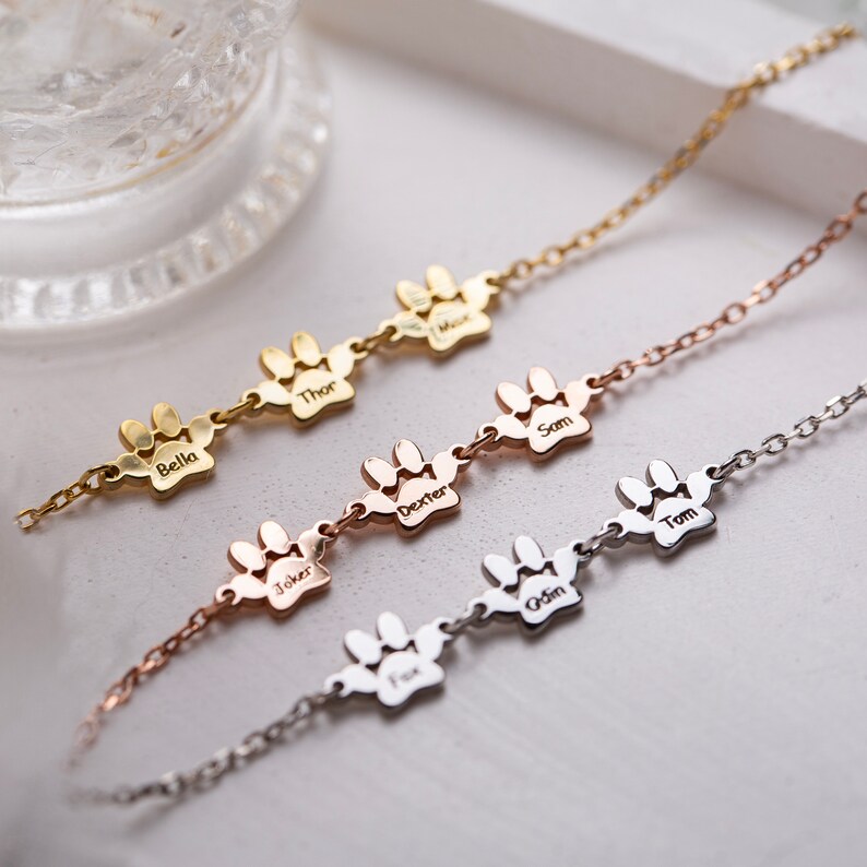 May include: Three gold, rose gold, and silver necklaces with paw print charms. Each charm is engraved with a different name: Bella, Thor, Dexter, Sam, Tom, Joker, Odin, and Finn.