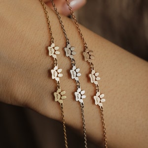 May include: Three gold chain bracelets with paw print charms. Each charm has a different name engraved on it: Bella, Thor, Max, Fox, Odin, Tom, Walker, Dexter, Sam.