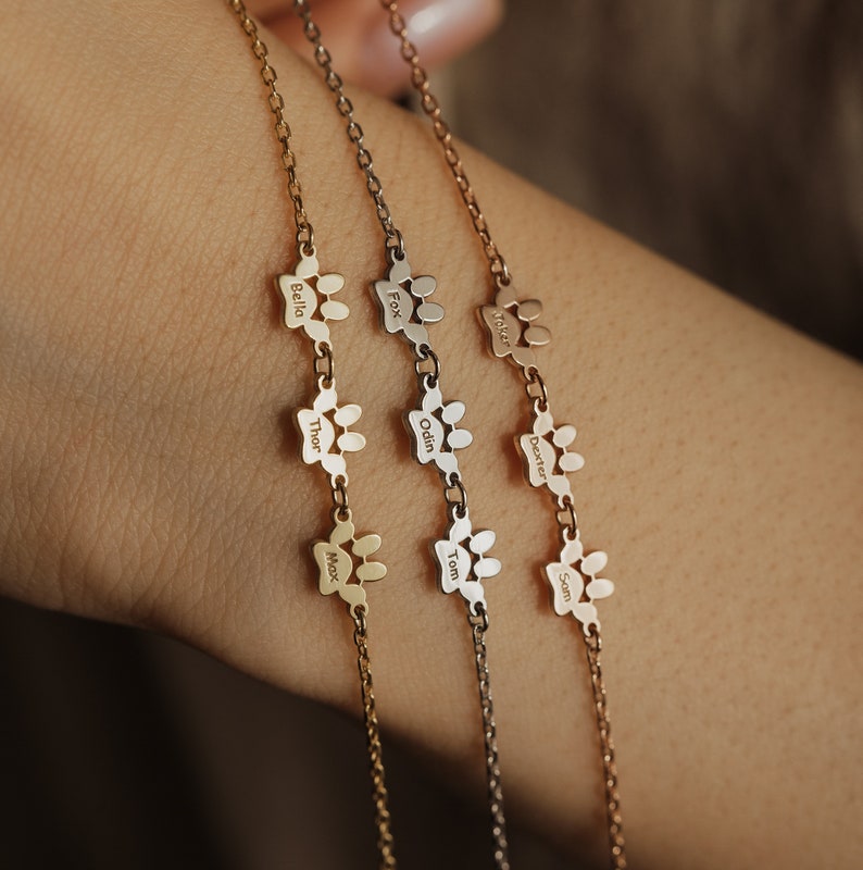 May include: Three gold chain bracelets with paw print charms. Each charm has a different name engraved on it: Bella, Thor, Max, Fox, Odin, Tom, Walker, Dexter, Sam.