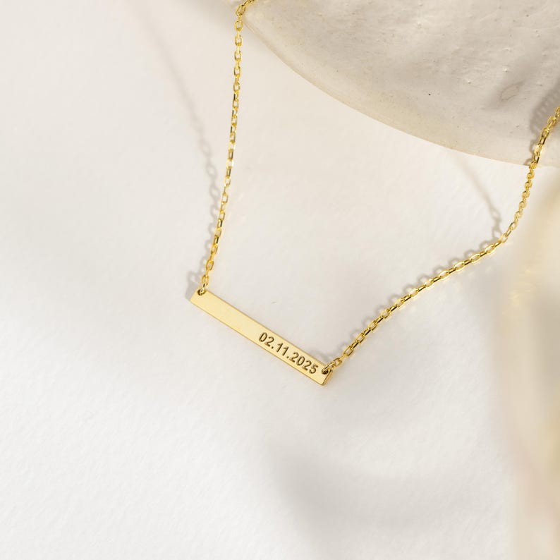 May include: A gold-colored bar necklace with a delicate chain. The rectangular bar is engraved with the date "02.11.2025". The necklace is displayed on a white surface, highlighting its simple elegance.