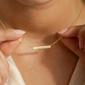 May include: A gold-colored necklace with a rectangular pendant. The pendant has the engraved date "02.11.2025". The necklace is held by a person. The background is a soft, neutral color.