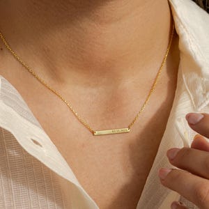 May include: A delicate gold-colored necklace with a rectangular pendant. The pendant has the engraved date "02.11.2025". The necklace is worn against a cream-colored shirt.
