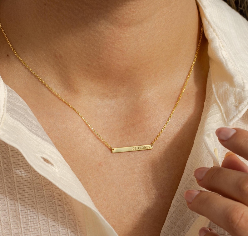 May include: A delicate gold-colored necklace with a rectangular pendant. The pendant has the engraved date "02.11.2025". The necklace is worn against a cream-colored shirt.