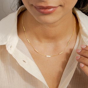 May include: A delicate gold necklace featuring small, rectangular bar pendants evenly spaced along a fine chain. The necklace is displayed on a person wearing a cream-colored button-down shirt. The jewelry is simple and elegant.