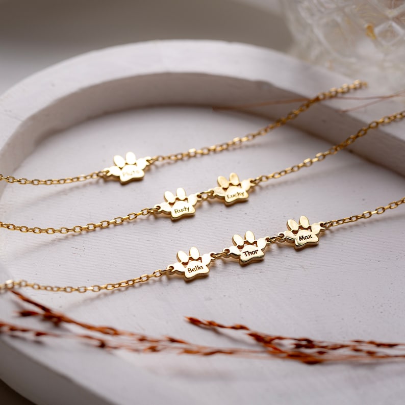 May include: Gold paw print necklaces with the names "Bella", "Thor", "Max", "Lucky", and "Rusty" engraved on them.