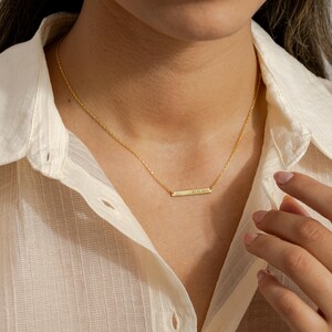 May include: A delicate gold-colored necklace with a rectangular pendant. The pendant has engraved text. The necklace is worn against a cream-colored button-down shirt. The necklace is a simple, elegant piece of jewelry.