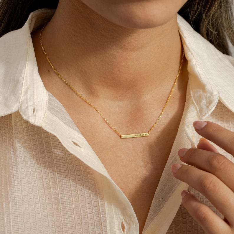 May include: A delicate gold-colored necklace with a rectangular pendant. The pendant has engraved text. The necklace is worn against a cream-colored button-down shirt. The necklace is a simple, elegant piece of jewelry.