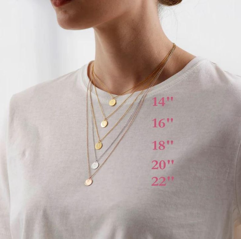 May include: A selection of layered necklaces with circular pendants in gold and silver tones. The necklaces are displayed on a white t-shirt, with pink text indicating lengths of 14", 16", 18", 20", and 22".