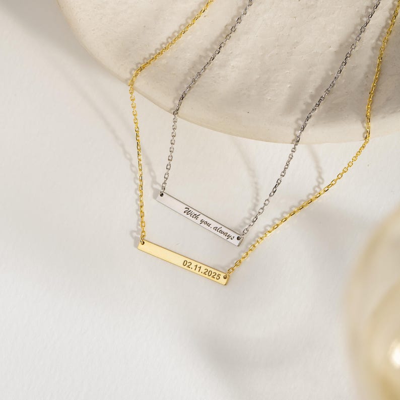May include: Two delicate necklaces with bar pendants. One is gold-toned with the date "02.11.2025" engraved. The other is silver-toned, inscribed with "With you, always." Both necklaces have fine chains and are displayed on a light background.