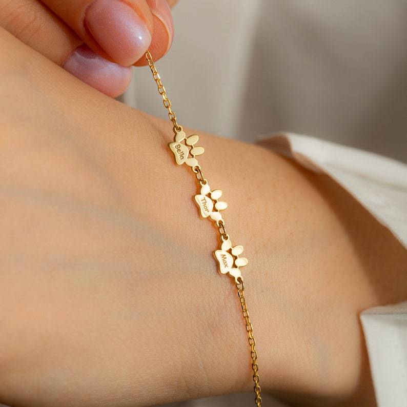 May include: Gold chain bracelet with three paw print charms, each engraved with a different name: Bella, Thor, and Max.