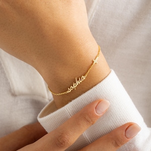 May include: A delicate gold bracelet with the name "Sophia" in cursive script. A small cross charm is attached. The bracelet is worn on a wrist, showcasing the jewelry's design and detail.