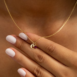 May include: A delicate gold necklace featuring a small letter "e" pendant and a rectangular ruby gemstone charm. The necklace is displayed against a skin tone background, highlighting the jewelry's fine details and elegant design.
