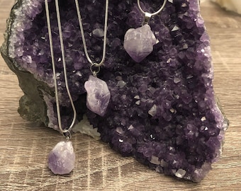May include: Three silver chain necklaces with amethyst crystal pendants. The necklaces are displayed on a purple amethyst geode.