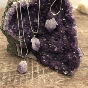 Puede incluir: Tres collares de cadena plateada con colgantes de cristal de amatista. Los collares se muestran en una geoda de amatista púrpura.