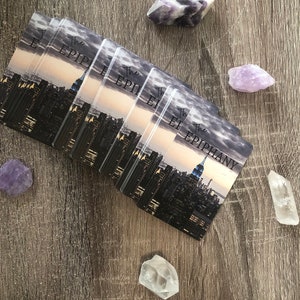 May include: A stack of cards with a cityscape background and the text "The Epiphany" on the top card. The cards are on a wooden surface with purple and clear crystals.
