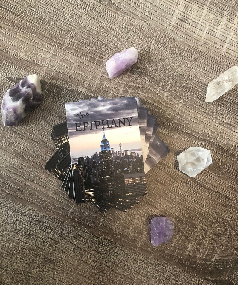 May include: A deck of cards with a cityscape and the words "The Epiphany" on the front. The cards are surrounded by purple and clear crystals on a wood surface.