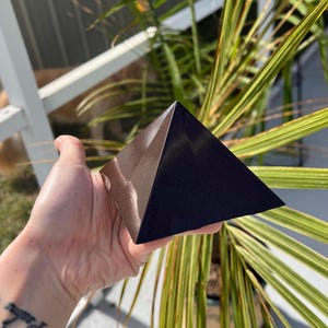 May include: A solid, smooth, dark brown pyramid object with a polished finish. The pyramid features four triangular faces meeting at a sharp apex, displaying a deep, uniform color with subtle light reflections on its angled surfaces.