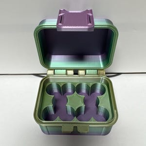 Könnte beinhalten: Eine quadratische, offene Aufbewahrungsbox mit Klappdeckel und Verschluss, die ein Farbverlaufsmuster von metallischem Grün zu tiefem Violett aufweist. Der Innenboden enthält einen geformten Einsatz mit vier kreisförmigen Aussparungen.